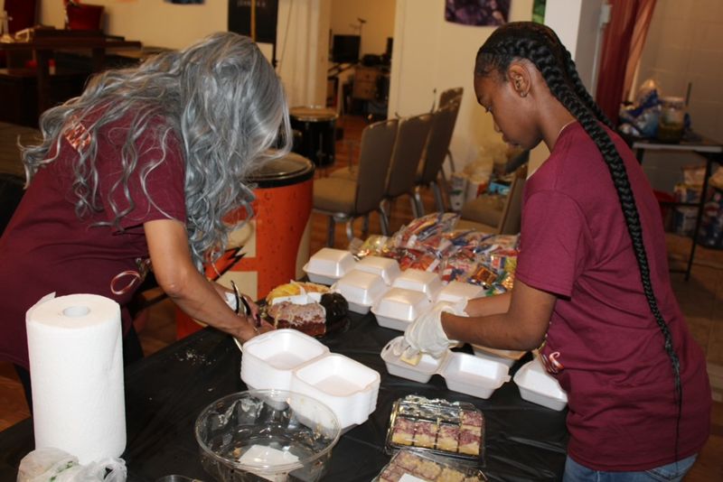Two volunteers at a serving table filling takeout containers with hot food.