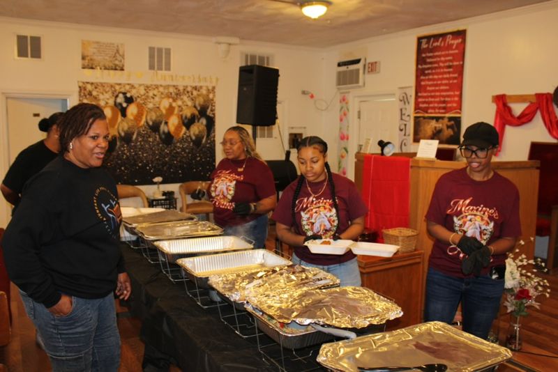 Volunteers standing behind a serving line of foil-covered trays in a church hall.