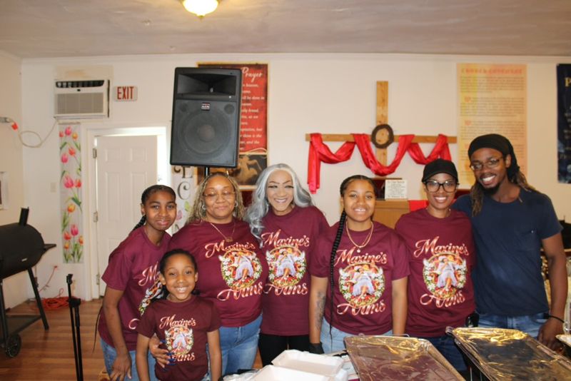 Seven volunteers of all ages in Maxine’s Arms shirts posing together in front of a decorated hall.