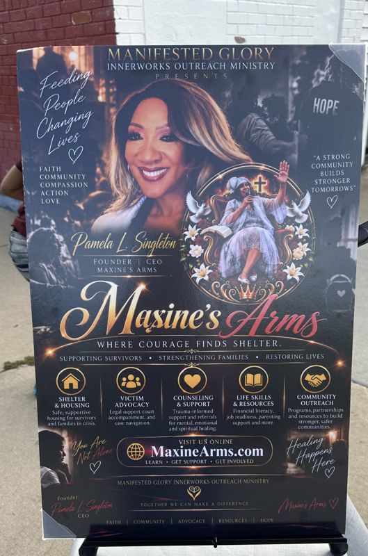 A tall Maxine’s Arms event banner on a sidewalk reading “Feeding People, Changing Lives” and listing shelter, advocacy, counseling, life-skills and community outreach services.